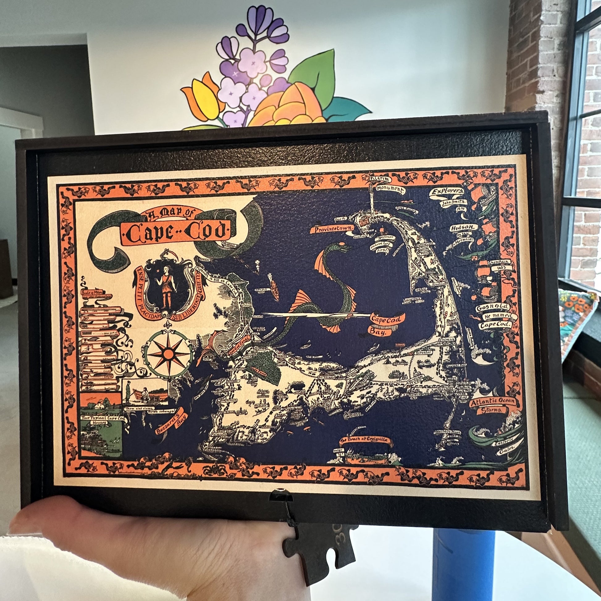 A hand holding a wooden jigsaw puzzle box of a vintage pictorial map of Cape Cod showcasing landmarks, natural wonders, cultural elements, and historical references. Illustrated cartography highlighting the state's diverse attractions and heritage. Artwork by Melanie Elisabeth and wooden jigsaw puzzle by WAWW Puzzles