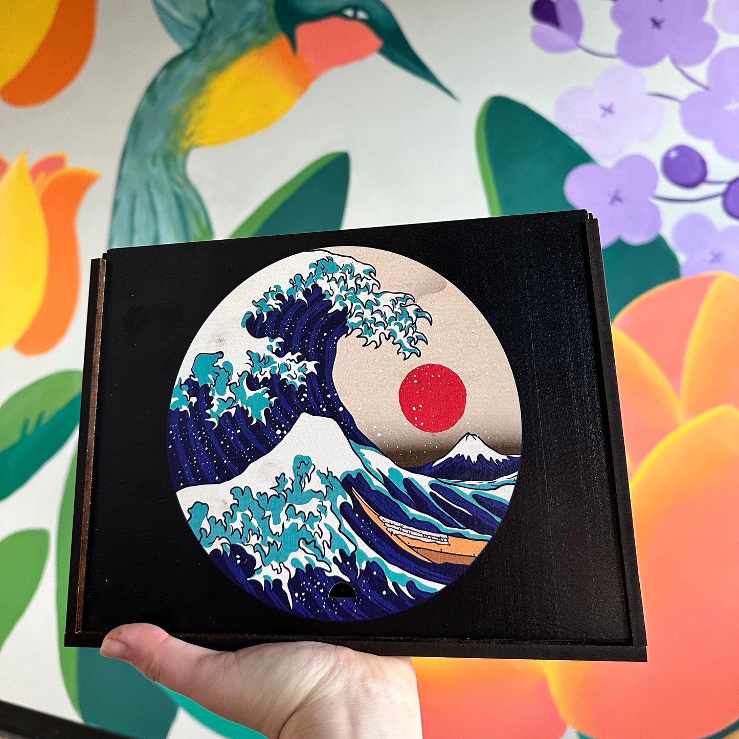 A hand holding a Handcrafted wooden box containing a colorful Japanese art jigsaw, similar to the Great Wave of Kanagawa intricate design.