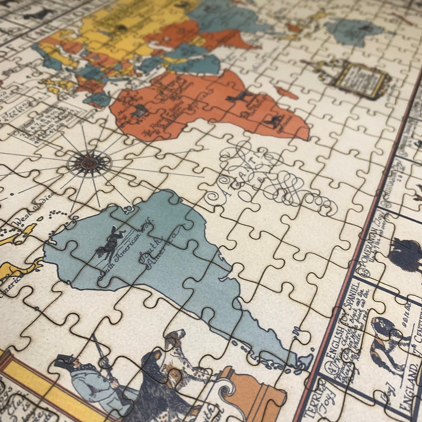 A close-up of a completed vintage-style world map jigsaw puzzle, showing colorful continents on an aged cream background with illustrated figures and decorative text.